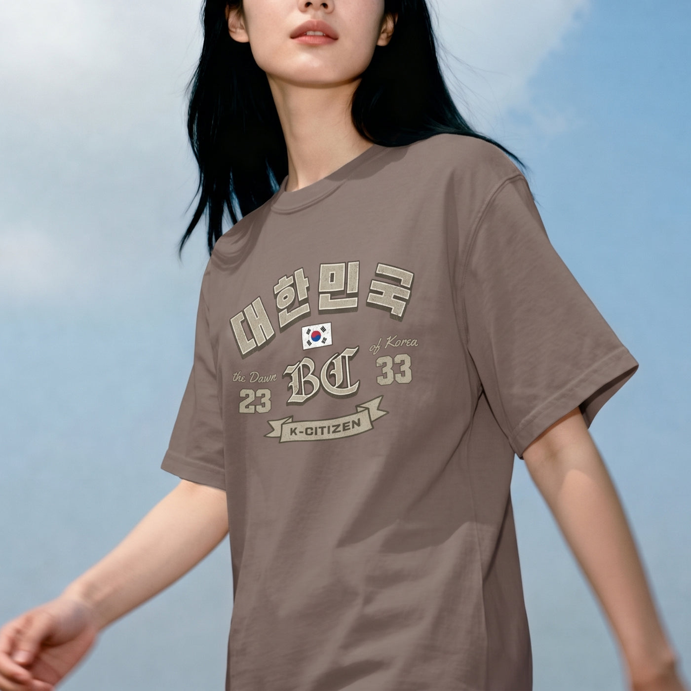 Lifestyle shot of a woman wearing a brown K-citizen t-shirt with a vintage collegiate Korean Hangul "대한민국" (Daehanminguk) typo