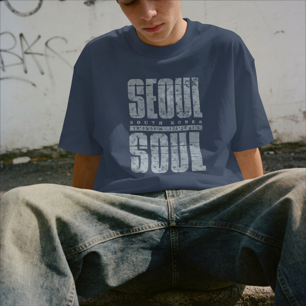 Person wearing a navy blue t-shirt with 'SEOUL SOUTH KOREA' printed on it, sitting against a graffiti-covered wall.