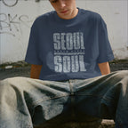 Person wearing a navy blue t-shirt with 'SEOUL SOUTH KOREA' printed on it, sitting against a graffiti-covered wall.
