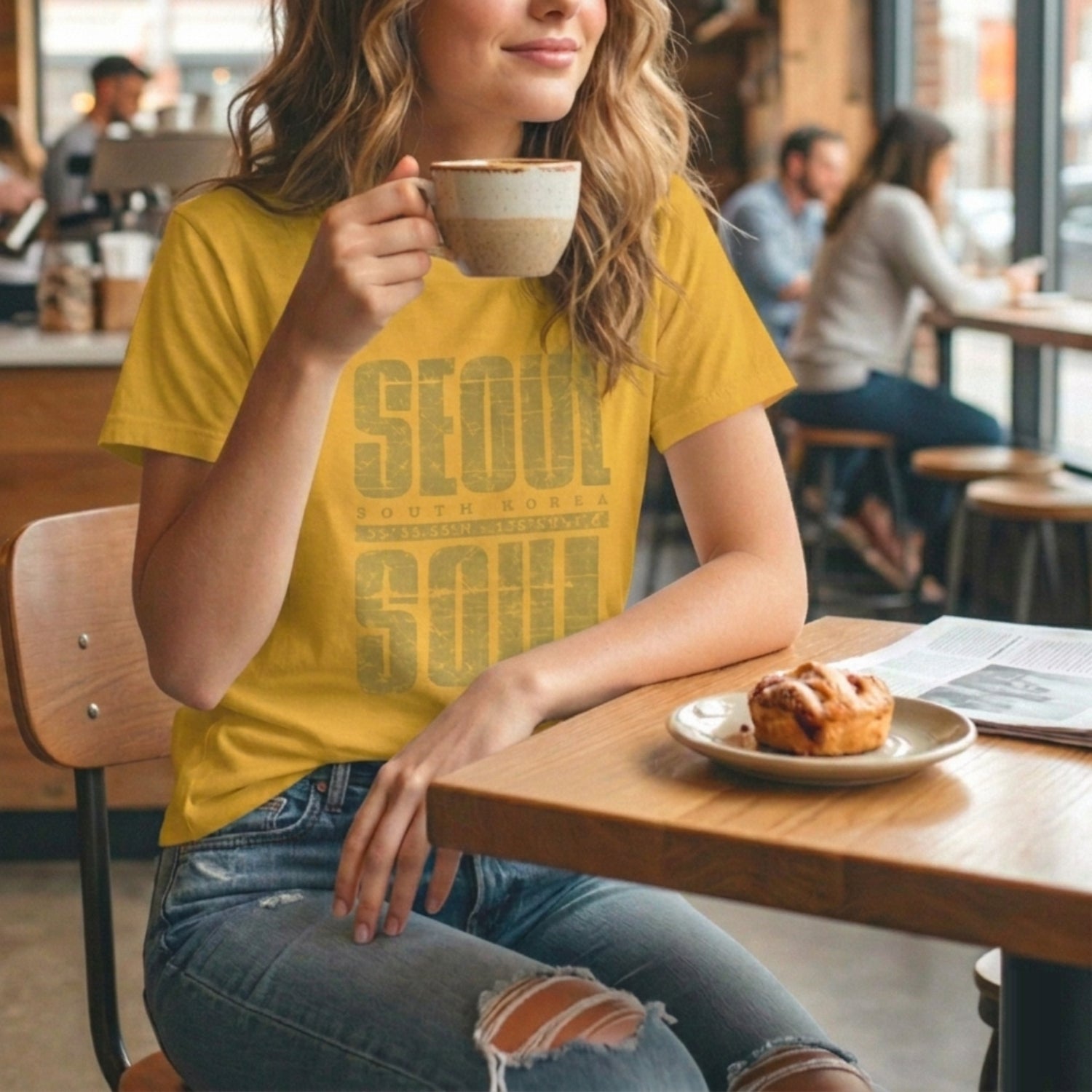 Lifestyle shot of a woman in a mustard yellow K-citizen t-shirt holding a coffee cup with Seoul Soul graphic