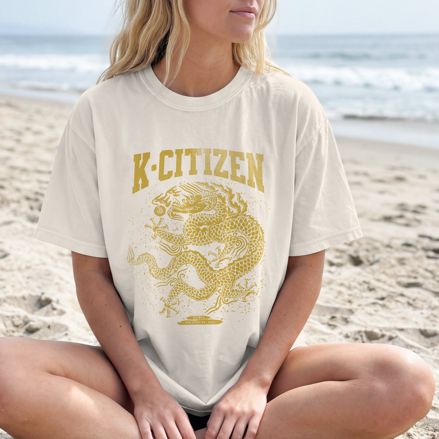 Person wearing a white t-shirt with 'K-Citizen' and a dragon graphic on a beach.