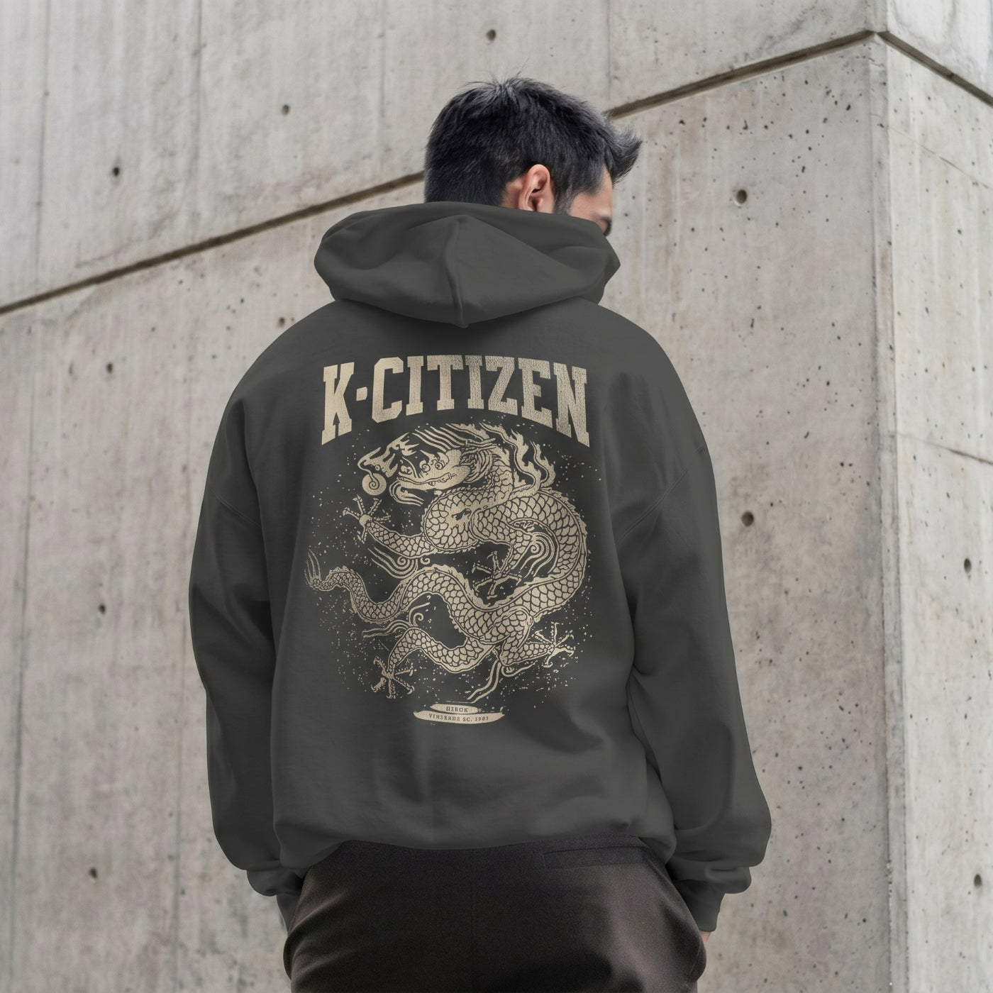 Person wearing a hoodie with a dragon design and 'K-Citizen' text on the back, standing against a concrete wall.