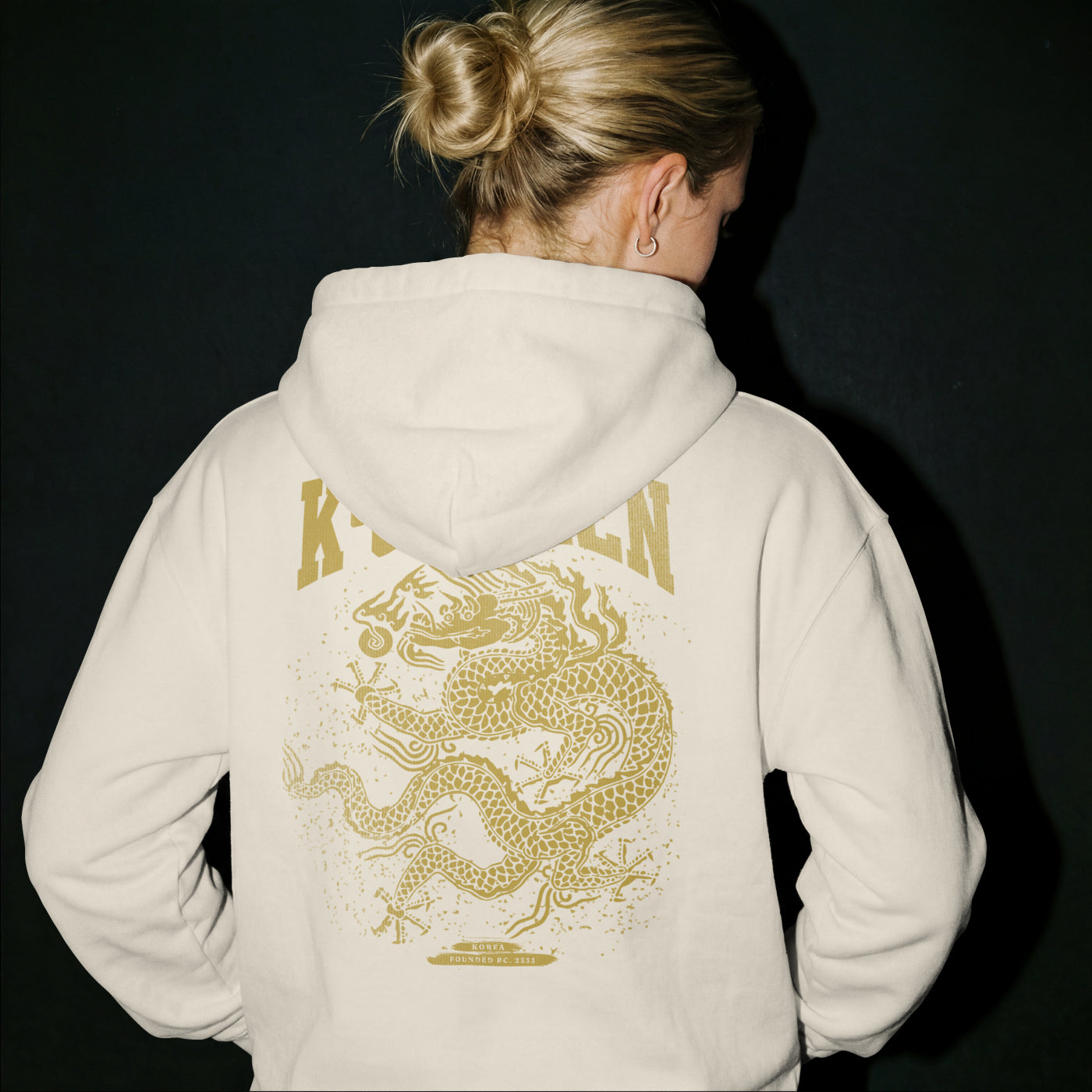 Person wearing a beige hoodie with a gold dragon design on the back against a black background